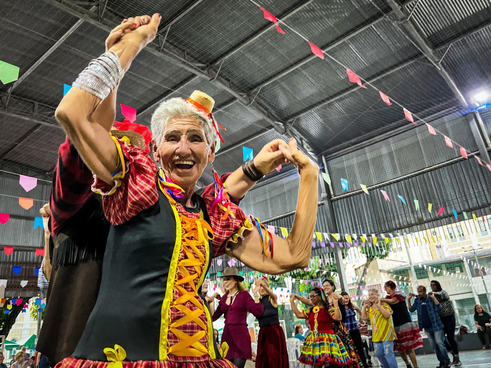 Idosa dançando sorridente em uma festa junina, com o grupo de pacientes e a equipe ao fundo.